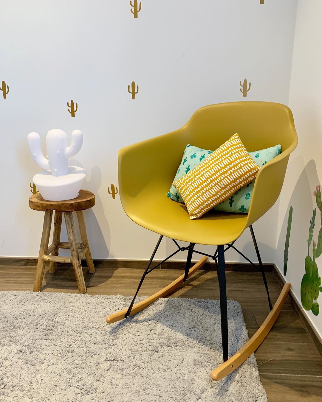 Rocking Chair Avon de couleur moutarde avec deux coussins rectangulaires; un jaune avec des points blancs et un vert d'eau avec des cactus. Il est situ&eacute; dans l'angle d'une piece dont les murs sont blancs avec des petits stickers de castus dor&eacute;s sur le mur de gauche et un gros de couleur verte sur le mur de droite. A gauche du rocking chair se trouve un petit tabouret en bois avec un cactus blanc pos&eacute; dessus. le sol est un parquent en bois recouvert d'un tapis gris/beige.